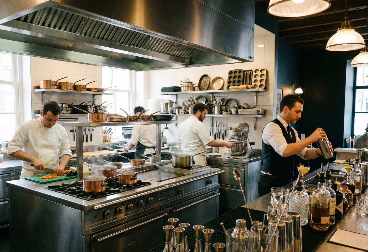 Bilden fångar en levande och autentisk ögonblicksbild från en högklassig restaurangmiljö där kök och bar möts. Den visar tydligt bredden i sortimentet genom att inkludera både matlagning och drinkblandning i samma scen. Detaljer: Köket (Vänster & Mitten Professionellt restaurangkök och bar i full aktivitet. Kockar förbereder mat vid en rostfri köksö med kopparkastruller medan en bartender skakar en drink omgiven av barutrustning.